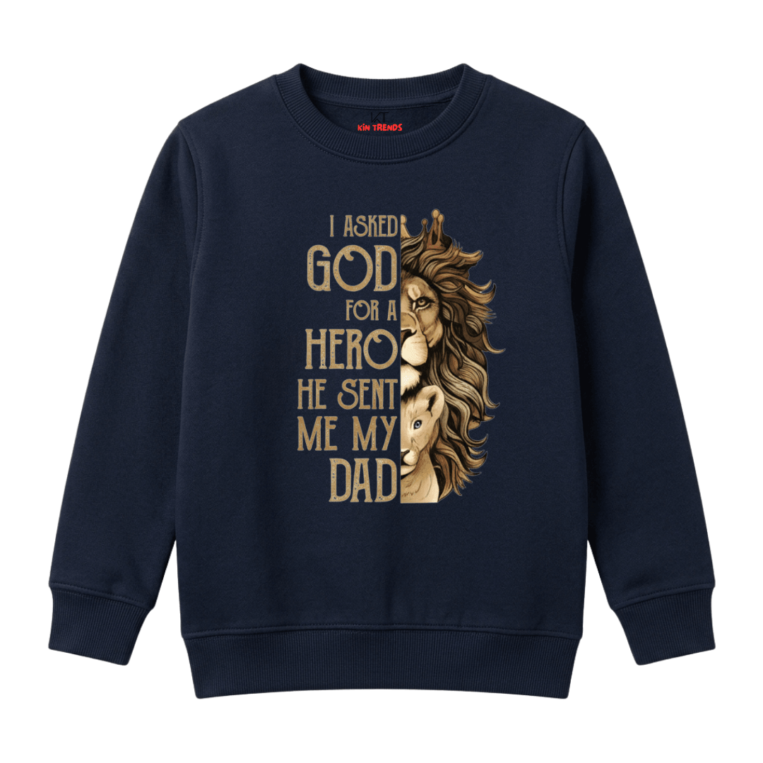 A navy blue crewneck sweatshirt featuring a graphic print of a lion half-severed from its human body, with the text 'I ASKED GOD FOR A HERO HE SENT ME MY DAD' in a gold serif font.